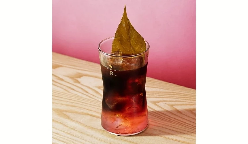 一杯櫻花桶陳冷萃咖啡放在木桌上,飲品呈現深咖啡與粉紅漸層,上方以櫻葉裝飾,玻璃杯印有星巴克臻選標誌
