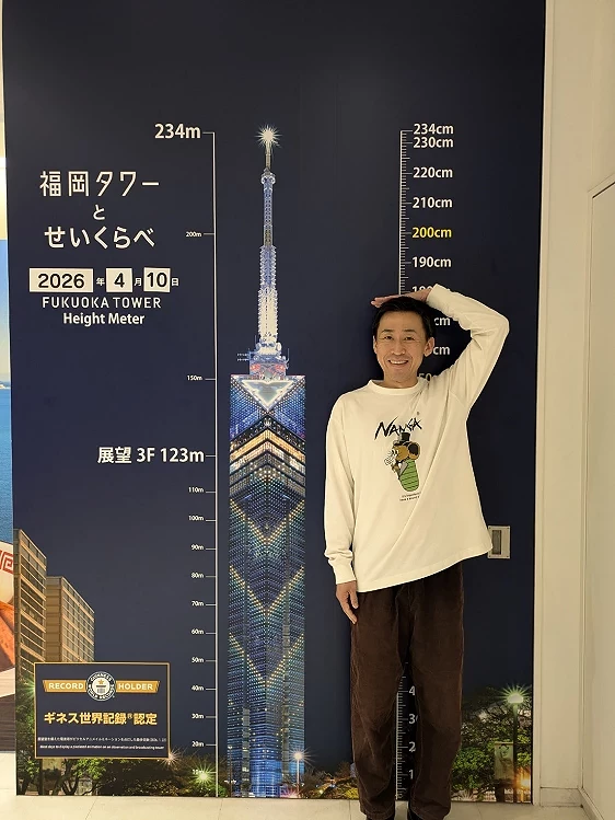 男性站在福岡塔的背板旁示意著身高的高度
