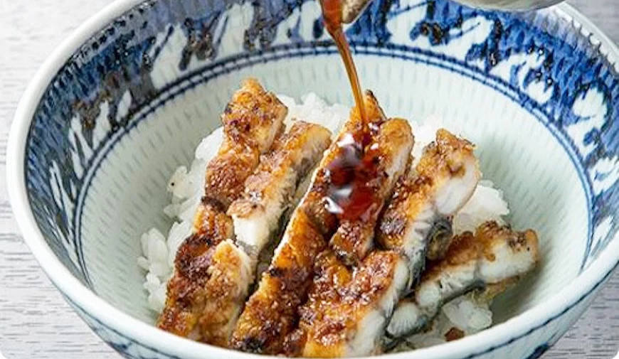 手持陶壺將醬汁淋於烤鰻魚飯碗,藍白花紋碗盛裝,魚肉表面呈焦褐色