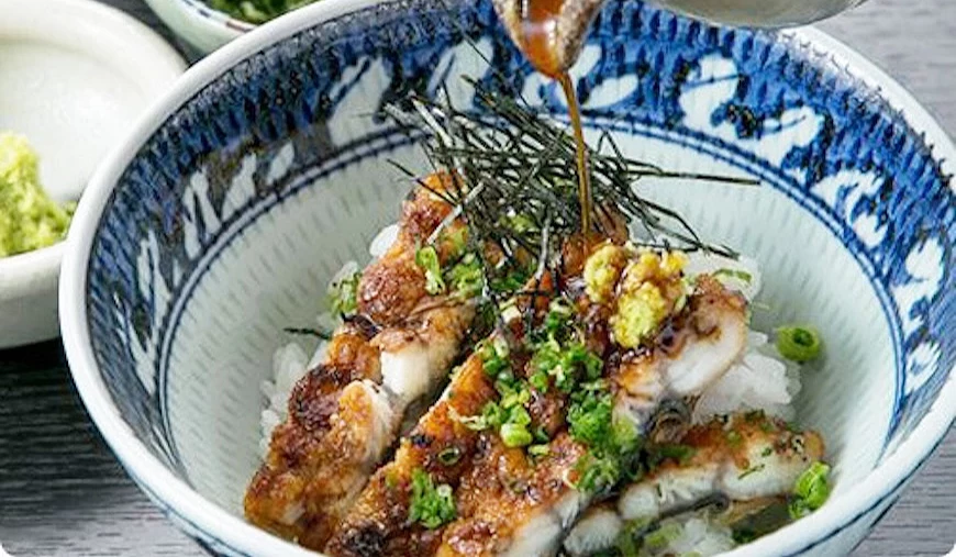 手持陶壺將醬汁淋於鰻魚飯碗,碗旁附有青蔥與山葵小碟,藍白花紋碗盛裝