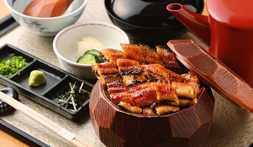 鰻魚飯盛裝於木製圓盒並掀蓋,旁附薬味盤、小黃瓜碗與紅色急須壺