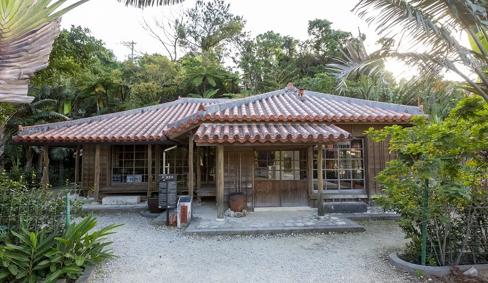建築為紅瓦坡頂、木造結構,設有寬敞的木格拉門與走廊,前方舖設石板路,門口右側屋脊上坐著一尊風獅爺。房屋周圍環繞著翠綠的亞熱帶植物與茂密樹林,展現出古樸且靜謐的琉球傳統農村風情。