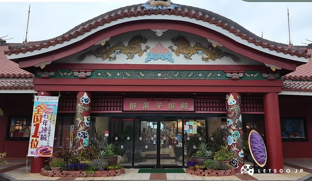「御菓子御殿 恩納店」的正門外觀照片。建築仿造首里城的門樓設計,擁有宏偉的朱紅色牆柱與琉球紅瓦屋頂,門額上飾有精緻的金龍浮雕,門柱兩側則有彩色龍紋圖騰。大門上方掛著寫有「御菓子御殿」的招牌,左側立有紅芋塔全國第一名的旗幟,右側則擺放著巨大的紅芋塔造型裝置。