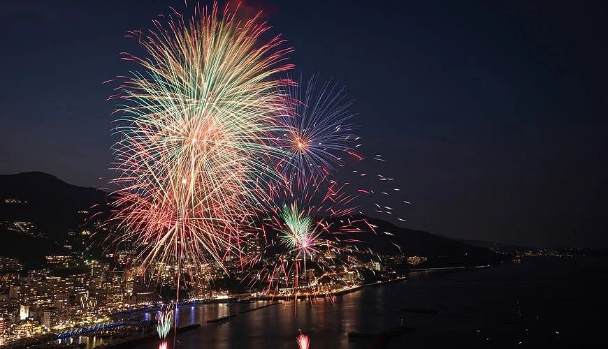 熱海海上花火大會於夜晚施放,五彩煙火在海灣上空綻放,下方可見熱海市區燈光與海岸線