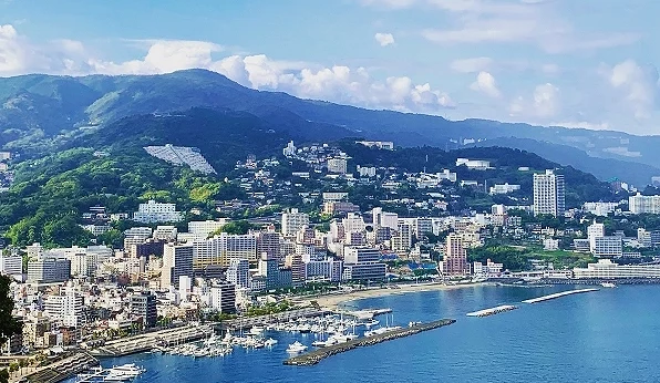 熱海市區與海岸線白天景色,沿岸可見飯店建築、港口與藍色海灣,背景為綠意山景