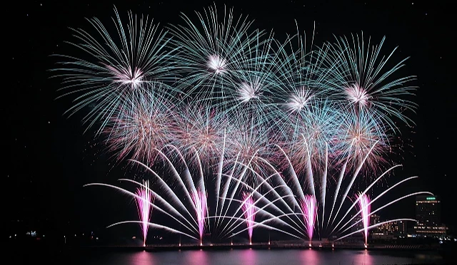 熱海海上花火大會夜晚煙火綻放,粉色、白色與藍綠色煙火照亮海灣上空,海面映出煙火光芒