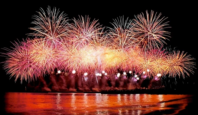 熱海海上花火大會於夜晚施放,大片金色與粉色煙火在海灣上空綻放,煙火光芒倒映在海面上