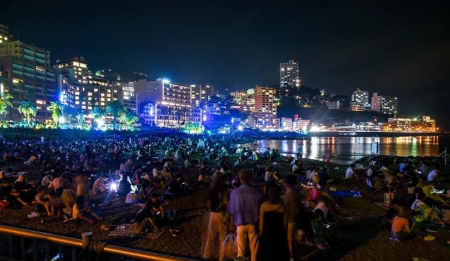 熱海花火大會夜晚會場人潮聚集,民眾坐在海灘上等待煙火施放,背景可見熱海市區與海灣夜景