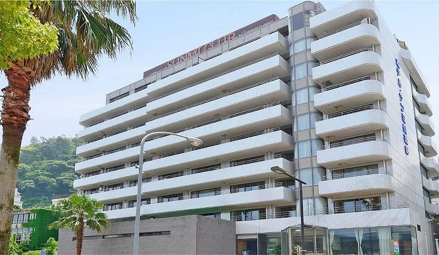 熱海海邊溫泉飯店外觀,白色建築設有多層陽台,前方有棕櫚樹,後方可見綠色山景