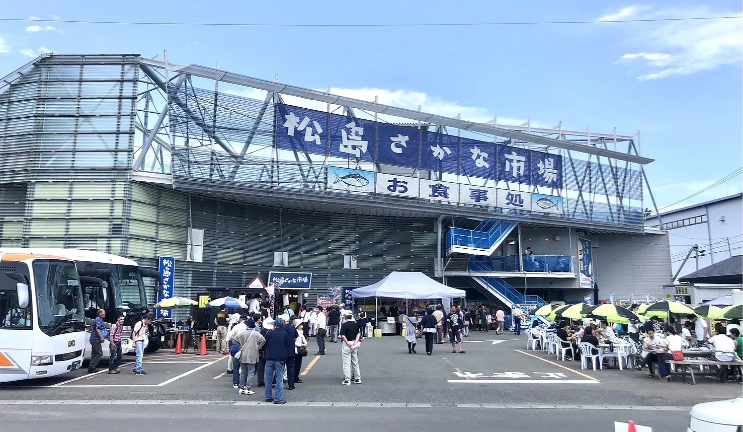 松島魚市場建築外觀,入口前有觀光客、巴士與戶外座位區,招牌寫著「松島さかな市場」。