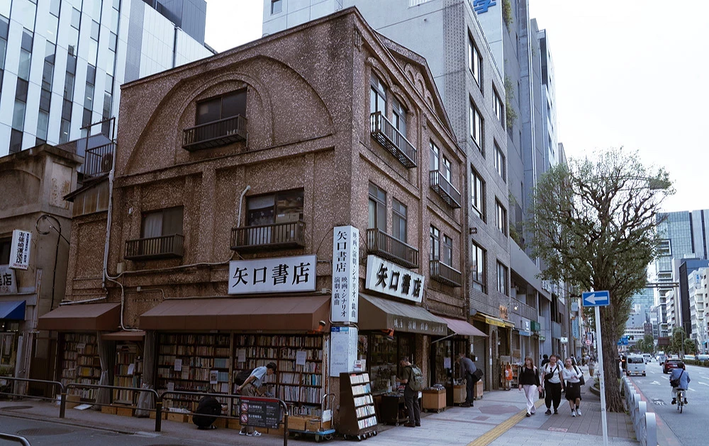 以矗立於轉角處的三層老木造建築為背景,展現出「矢口書店」白底黑字的大招牌與整面露天書牆,呈現該店面特有的懷舊街頭景觀特徵