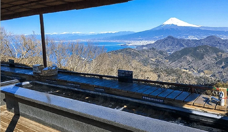 山頂足湯泡腳池全景,背景為積雪富士山、山脈與海灣,晴天藍天,有屋頂遮蔽