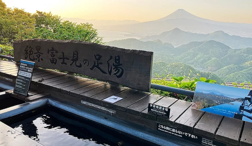 山頂露天足湯設施,木製看板寫有「絕景 富士見の足湯」,背景可見富士山與連綿山脈,夕陽西照
