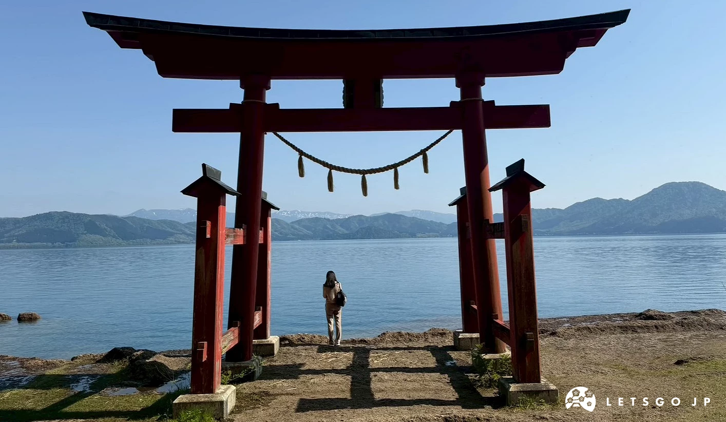 一名女子獨自站在面向平靜藍色湖面的紅色大鳥居下方,背景視野遼闊。