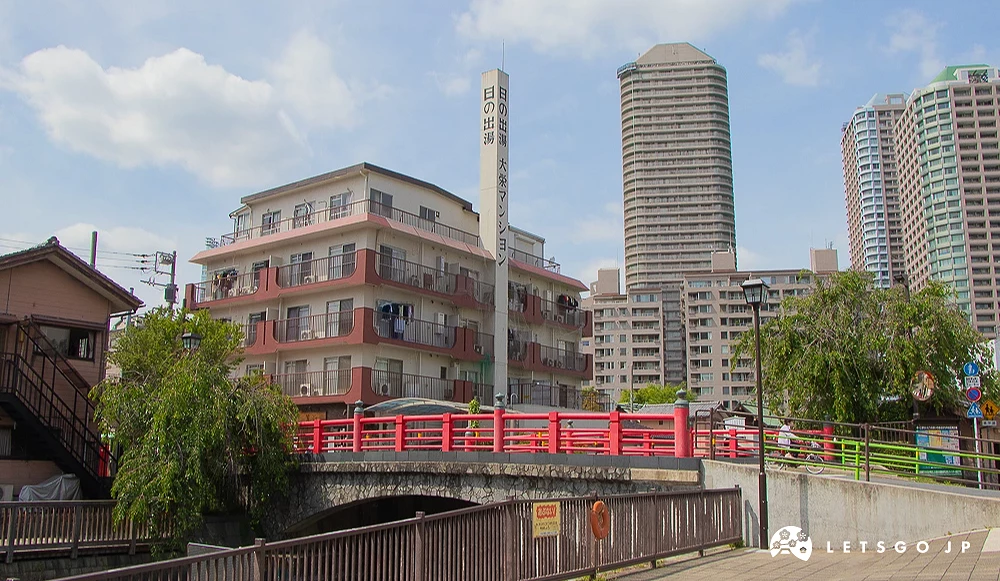 月島西仲橋與日の出湯 石造拱橋設有紅色欄杆,背景為寫有「日の出湯」的煙囪與高樓大廈。