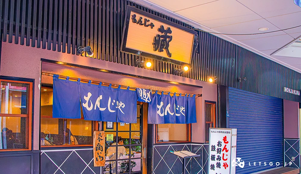文字燒藏店面外觀 店門口掛著深藍色「もんじゃ」暖簾,上方有木刻招牌,右側立著文字燒「藏」的燈箱。