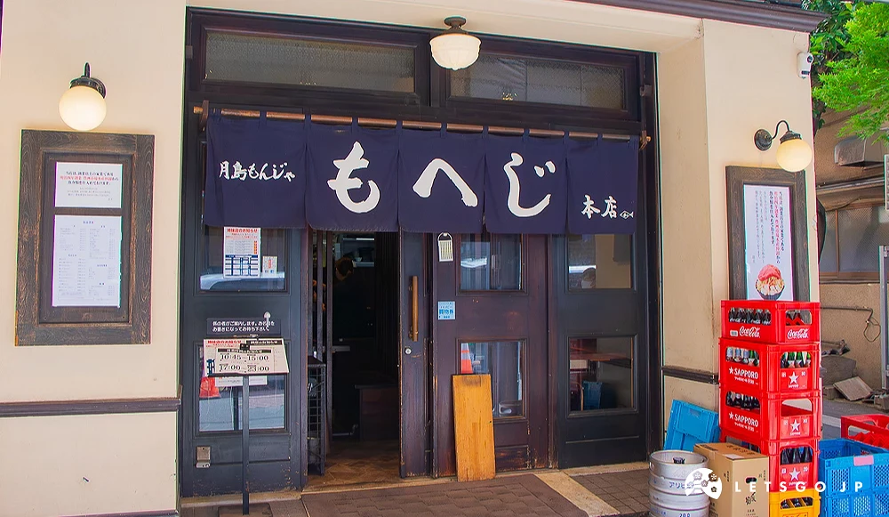 もへじ文字燒本店入口 深色木門上方掛著寫有「月島もんじゃ もへじ 本店」的藍色暖簾,門口放置酒籃與啤酒桶。