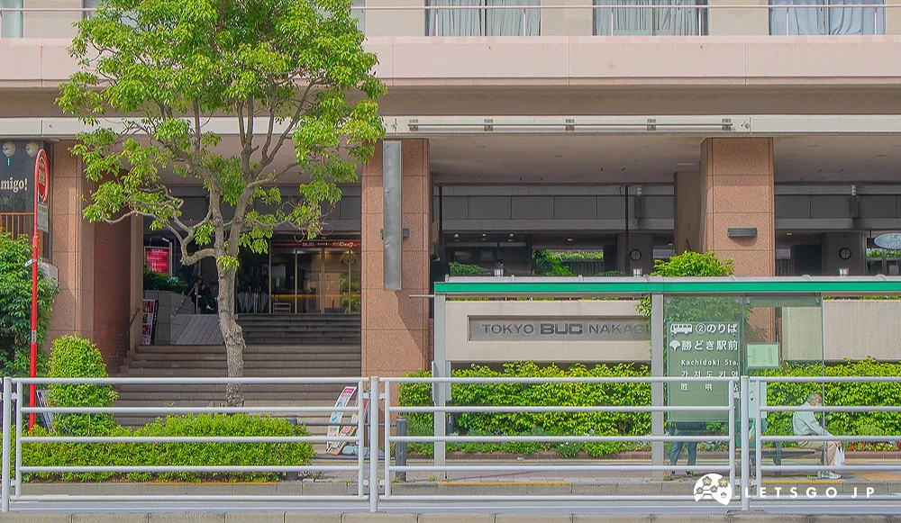 勝どき站前公車站 TOKYO BUC 建築前方設有綠頂公車站,站牌標示「勝どき駅前」,長椅上有一名旅客。