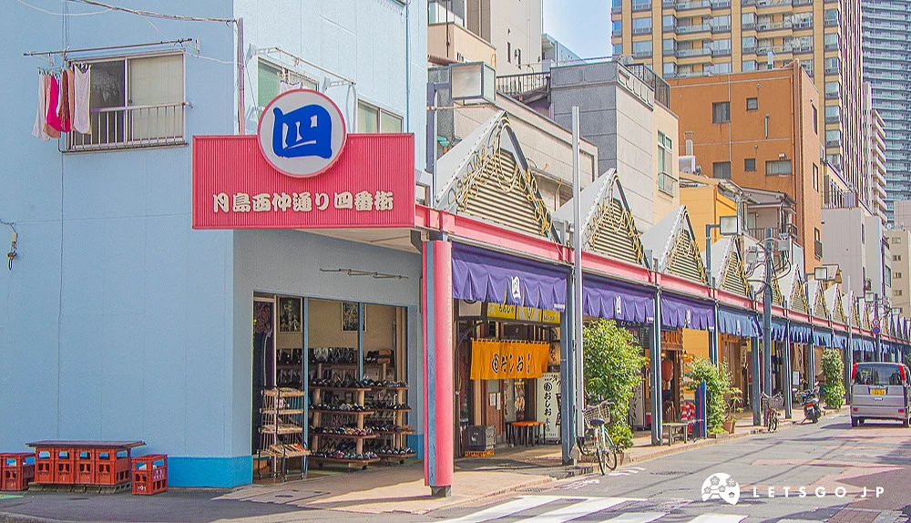 月島西仲通四番街入口 桃紅色拱門招牌上寫著「月島西仲通り 四番街」,右側延伸出帶有深藍色暖簾的商店街街景。