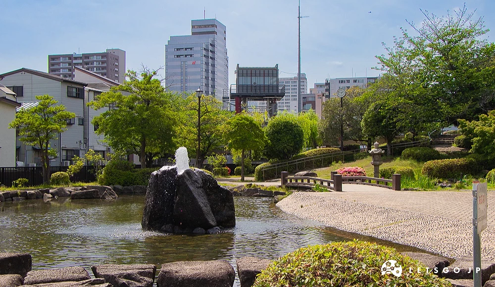 月島佃公園噴水池 佃公園內的池塘中央設有岩石噴水池,右側有木造小橋,後方為城市建築。