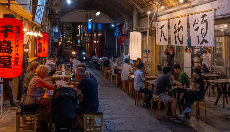一條街道上,左右兩側有許多居酒屋,有一群人坐在那裡吃飯喝酒