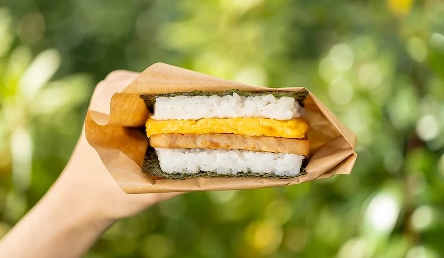 手持以海苔與白飯夾住煎蛋及豬排的飯糰三明治,以牛皮紙包覆,戶外綠意背景