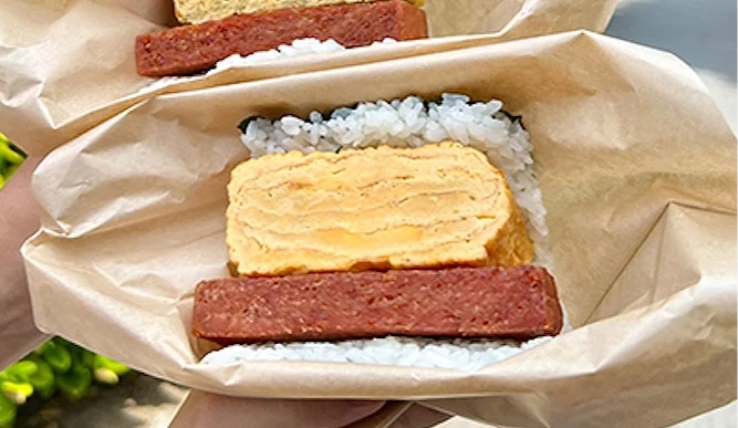 雙手各持一份以牛皮紙包覆的午餐肉玉子飯糰,戶外綠樹與道路為背景