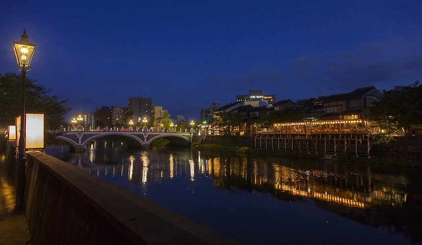 夜幕低垂河畔古鎮,石橋與木造建築燈火通明,倒影映於靜水景致優美。