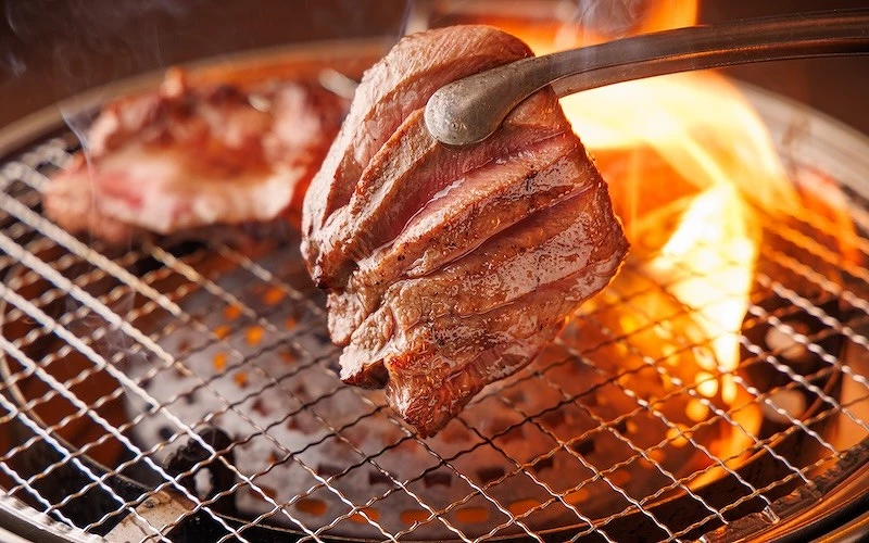 炭火烤爐上以夾子翻動厚切牛肉，火焰竄起