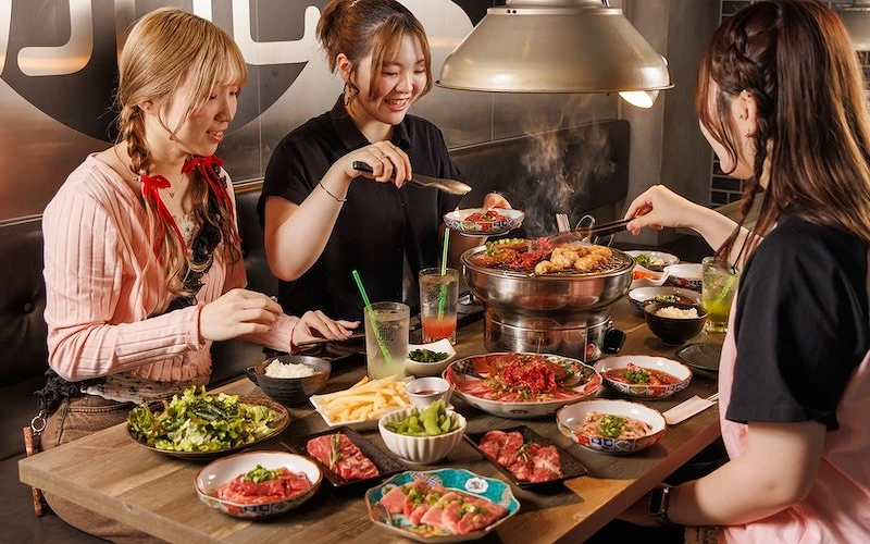 三位女性在燒肉店圍桌用餐，服務人員協助夾肉，桌上擺滿各式肉品