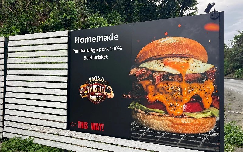 戶外看板印漢堡照片,標示「YAGAJI MUSCLE BURGER」與「Homemade Yambaru Agu pork 100% Beef Brisket」