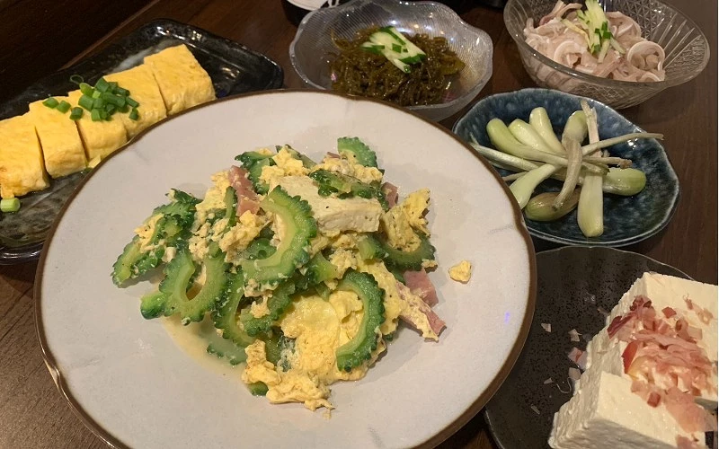 白盤盛苦瓜炒蛋,周圍有玉子燒與多樣小菜