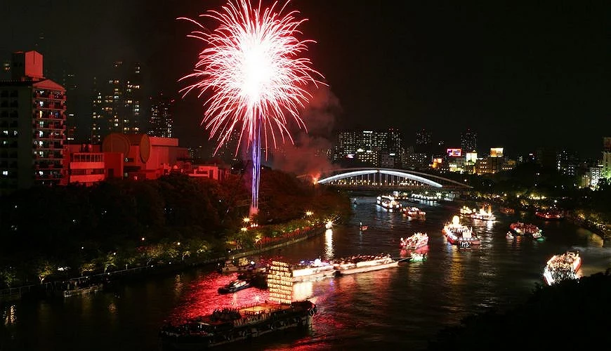 大阪天神祭奉納煙火於大川上空綻放,河面上可見船渡御船隊與燈火倒影,展現夏季祭典夜景
