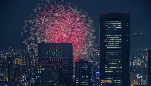 大阪市區高樓夜景間綻放的天神祭奉納煙火,大片紅色煙火點亮大阪夏夜天際線