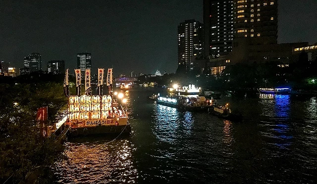 天神祭船渡御的船隊夜晚行經大阪大川,提燈燈火與周邊高樓夜景倒映在河面上