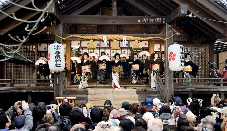 日式神社舞台表演,舞者穿著和服起舞,白色燈籠與繩飾點綴,遊客聚集觀賞祭典。