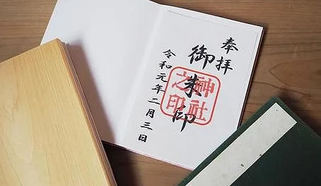 一本白色的書本頁面,上面用黑字寫著「奉拜 御朱印」,後面蓋有紅色的神社印章。