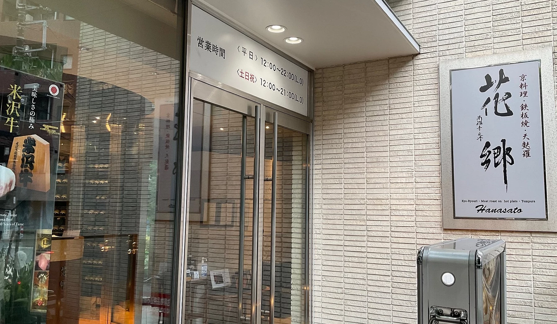 東京六本木花鄉餐廳的現代感大門入口,玻璃自動門旁貼有白色告示,右側米色磚牆上掛著寫有花鄉字樣的質感方形招牌。