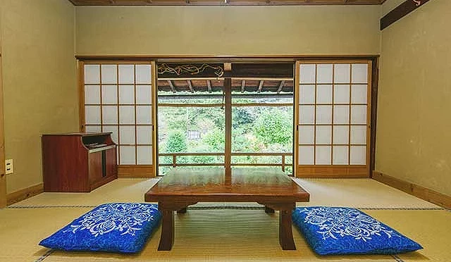 一個和式房間,裡面有一張桌子,兩邊有座椅墊。後面是日式拉門,拉門後面是綠色的景觀。