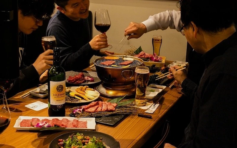四人圍坐享用燒肉，桌上擺有紅酒、啤酒、多盤生肉與銅製烤爐