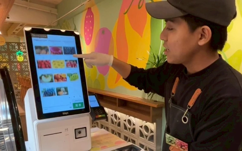 戴帽店員站在觸控點餐機旁,手指螢幕上的水果選單
