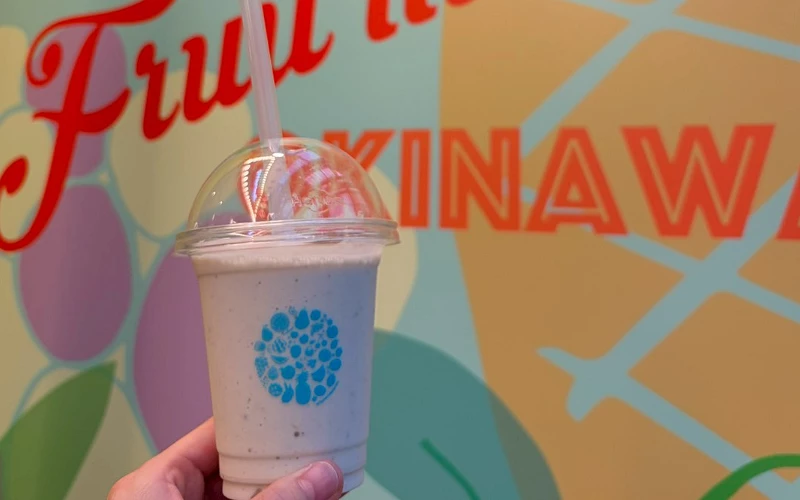 手拿一杯白色冰沙,背景牆印紅字與「OKINAWA」字樣