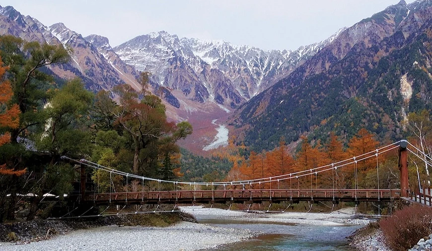 `懸索橋橫跨淺灘河流,背景為秋葉紅樹與積雪山峰`