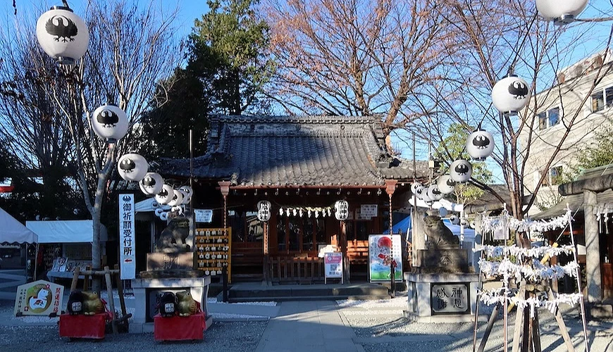 日本神社境內,白色燈籠掛於樹枝上,燈籠印有八咫烏圖案,正面為朱紅色拜殿,左側立有「御祈願受付所」指示牌,右側掛有繪馬與御守。