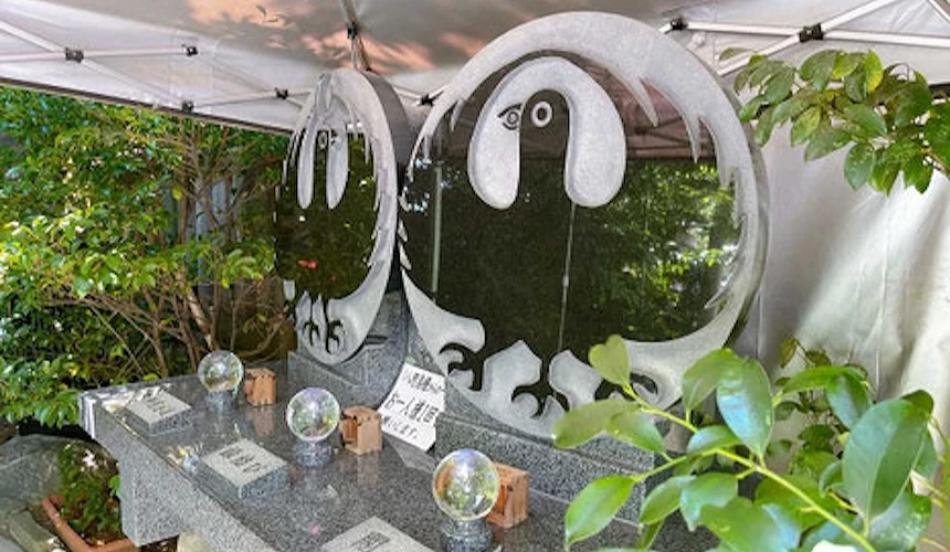 戶外帳篷下展示兩座圓形花崗岩墓碑,碑面刻有黑白動物圖案,前方石台上擺有水晶球與木製小物,寫有日文標語