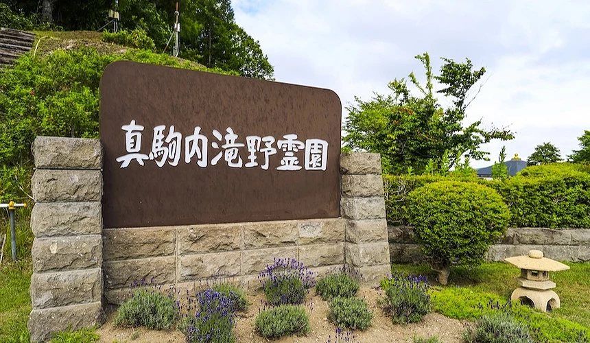 棕色石碑寫有日文標語，灰石柱基座與蒼翠樹林映襯，紫薰衣草與石燈籠點綴園區。