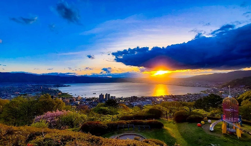 金色夕陽沉入湖面,城鎮與山景映照,暗藍雲彩與橙黃光芒,綠樹與紫紅花卉點綴前景,兒童遊樂設施靠近,日本湖畔黃昏風景絢麗壯闊。