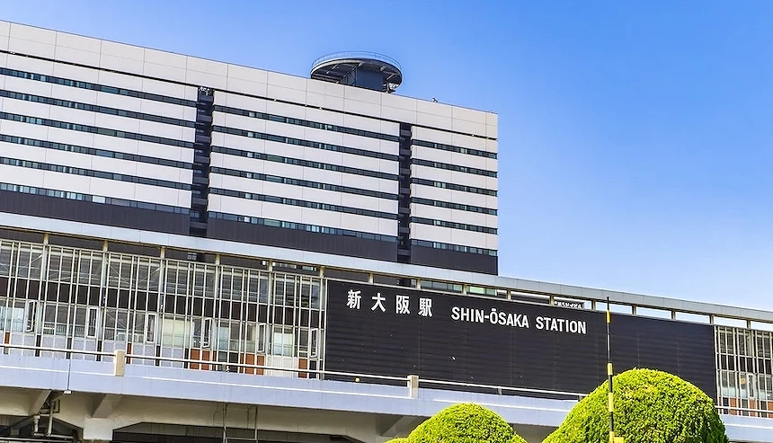 新大阪站現代建築立面,白黑條紋玻璃牆與圓形頂部,日文與英文標語清晰,綠灌木點綴。