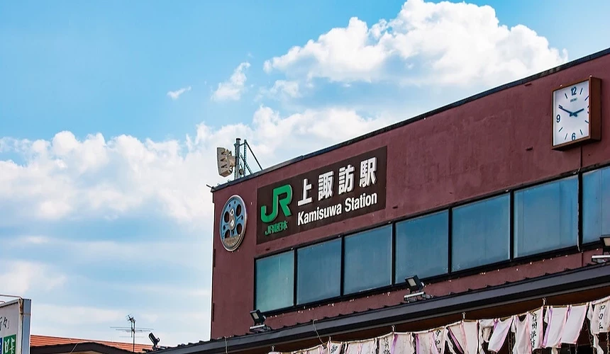 褐紅色車站建築,綠色日文站名標誌與英文Kamisuwa Station,白色時鐘與JR標誌,玻璃窗排列整齊,紫紅色布條與白色書法標籤懸掛屋簷,晴朗藍天與白色蓬鬆雲朵,日本鄉村火車站建築典型。