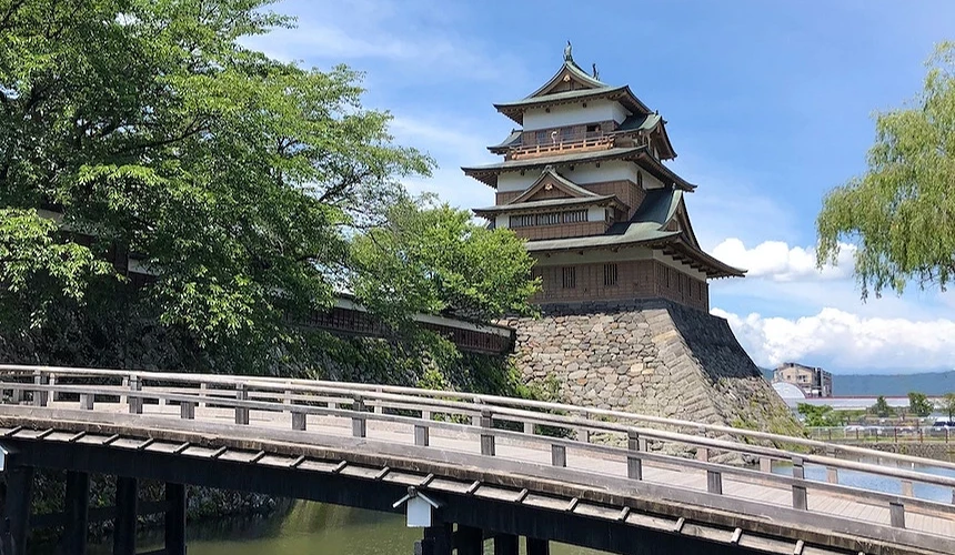 木製拱橋跨越護城河,白色欄杆與黑色橋樑,古城堡聳立背後,褐色木造多層翹角屋頂與灰白石牆基座,綠樹環繞兩側,遠景藍色湖水與城鎮,晴朗藍天映照,日本古城堡建築歷史悠久莊嚴宏偉。